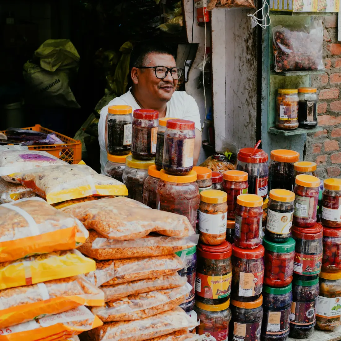 A smiling shopkeeper behind a counter stacked with jars of pickles and packets of snacks