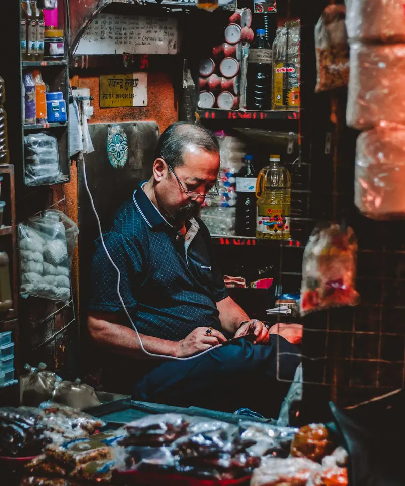 A shopkeeper in Kathmandu checking his phone at the counter of his small shop, shelves of goods around him