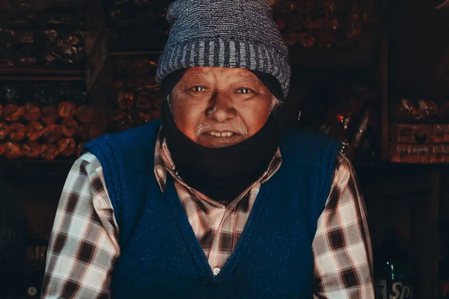 An elderly shopkeeper in a woollen cap leaning on the counter of his shop in Chitlang, Nepal