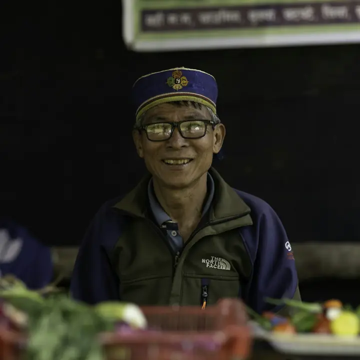 A smiling vegetable seller in a cap and glasses behind his stall near Kathmandu